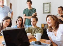 Mehrere junge Frauen und Männer vor Laptop um Tisch sitzend und stehend